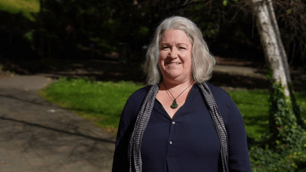 A grey-haired woman wearing a navy top and a pounamu stands on a lawn