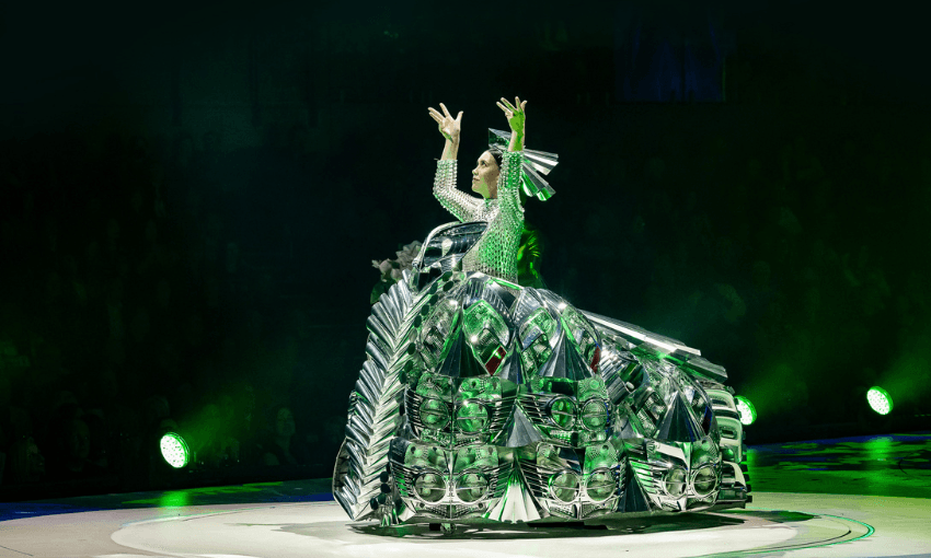 A performer in a shiny, futuristic silver costume with geometric patterns raises their arms on a circular stage, illuminated by green spotlights against a dark background.