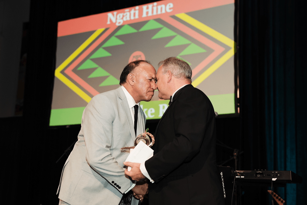 Two men share a hongi, a traditional Māori greeting, on stage in front of a screen displaying "Ngāti Hine" and a colourful geometric pattern. One holds a gift and an envelope.