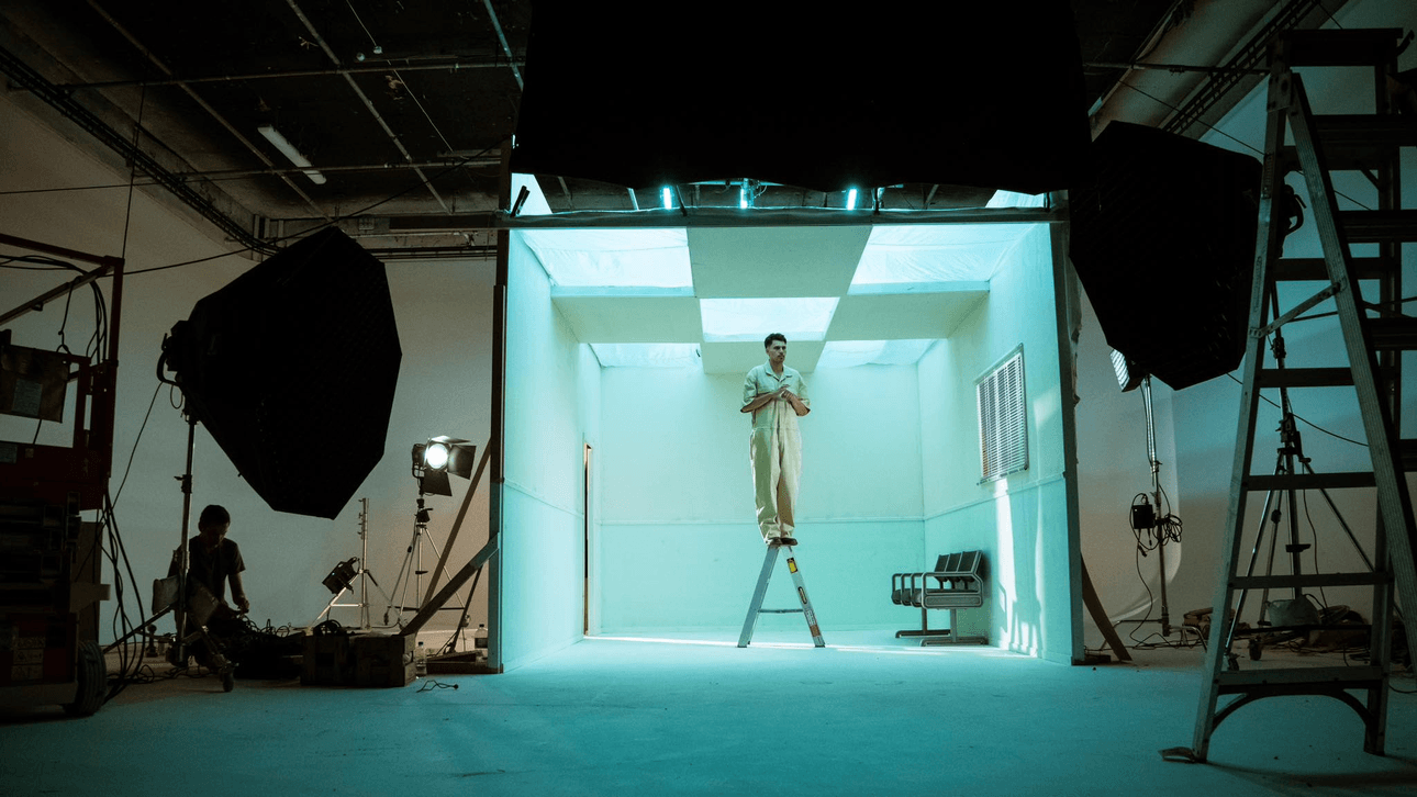 A person in light-coloured clothing stands on a ladder inside a brightly lit, blue-tinted room, surrounded by lighting equipment and ladders in a studio setting.