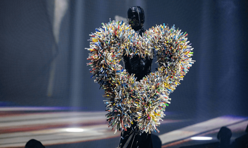 A person in a black, full-body suit stands on stage wearing a large heart-shaped costume made of colorful, densely packed objects, resembling pens or markers, with dramatic stage lighting in the background.