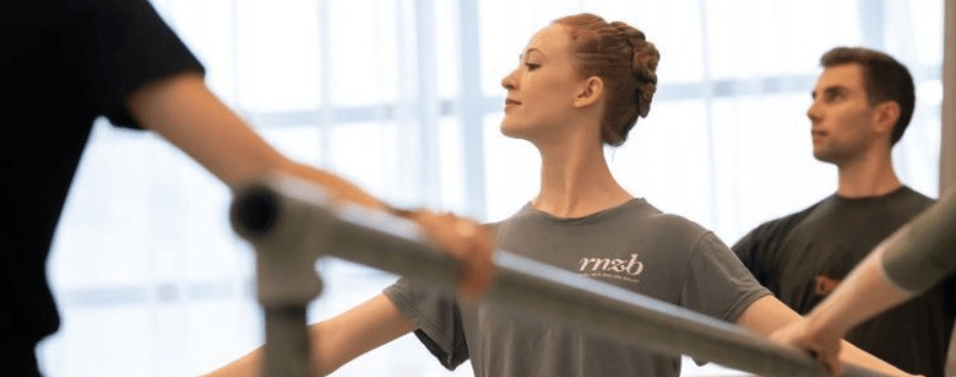 Three dancers are at a ballet barre. The image focuses on a young woman who has her hands spread and she is looking away from the camera.