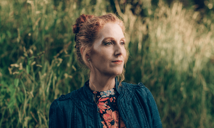 A photograph of Catherine Chidgey, the writer, who has red hair tied in a bun and is looking into the distance. A background of long grass is behind her.