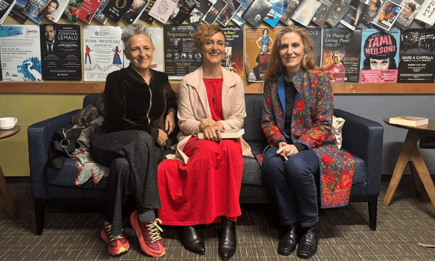 A photograph of three woman sitting side by side on a couch. They are all smiling. Behind them is a wall covered with posters. 