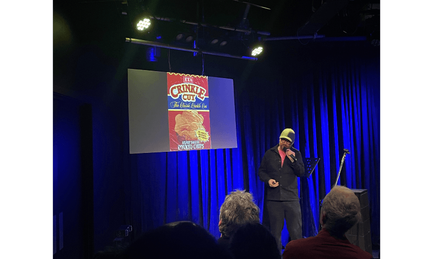 A photo of a stage washed in blue light. There is a screen showing a picture of an ETA chip packet "crinkle cut". There is a man, Duncan Sarkies, standing at a mircrophone.