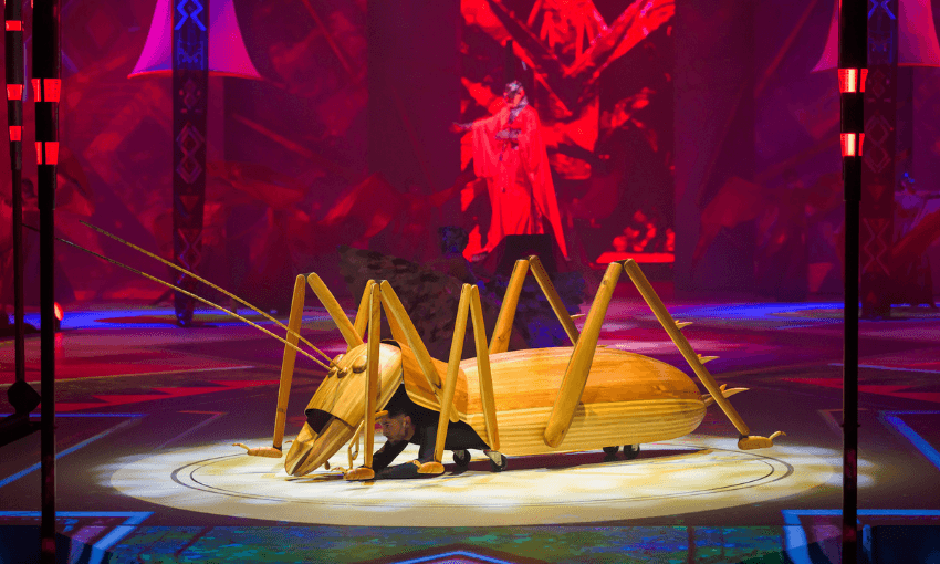 A performer in a large, stylized grasshopper costume crouches on a brightly lit circular stage, surrounded by vivid red and purple lighting and theatrical set pieces.