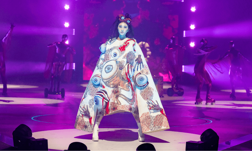 A model on a lit stage wears a dramatic, oversized cape decorated with surreal eye and hand designs. Performers in futuristic costumes and roller skates pose in the purple-lit background.