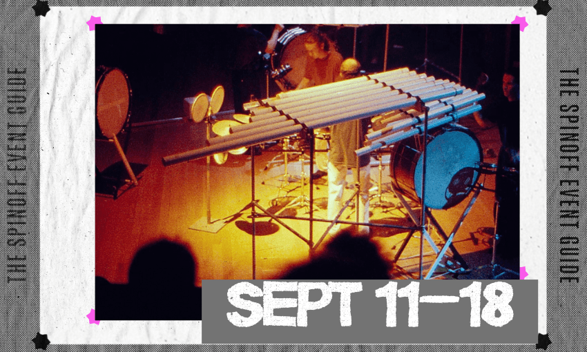 The image of a blurry stage with large instruments set up including a xylophone and several drum kits. Three musicians stand on stage, playing.