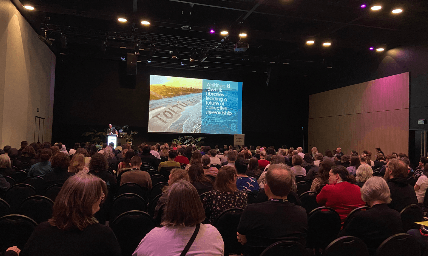 A photo of a large room with people seated. There is a stage at the front with a large screen. A person is standing at a lectern.