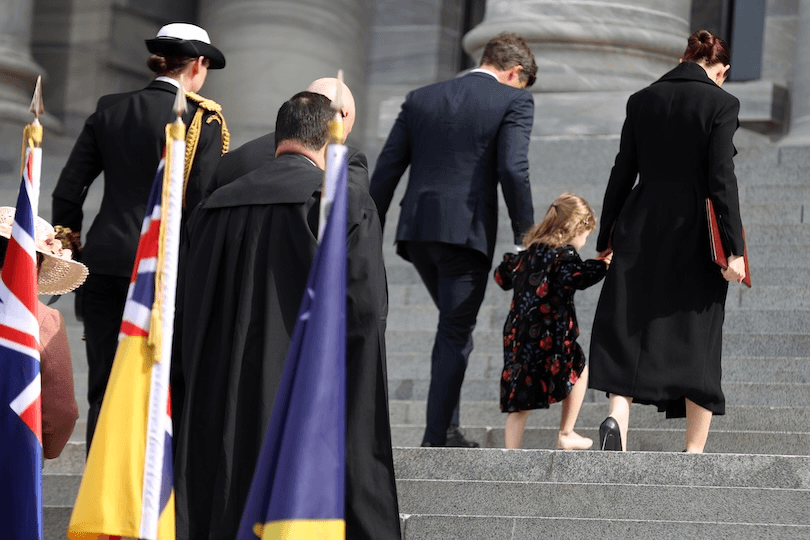 A group of adults and a young girl, seen from behind, walk up stone steps outside a grand building. Flags are in the foreground, and the child holds hands with a woman in a black coat.