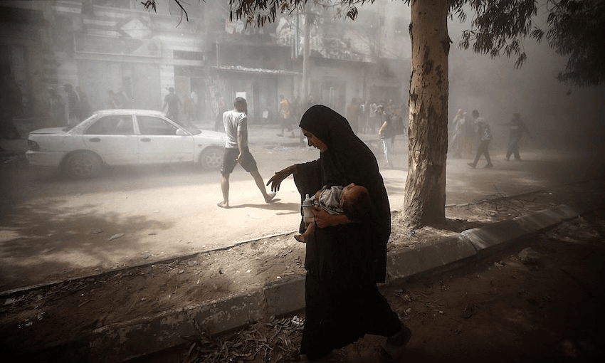 Palestinians watch as the Al-Ghafri tower collapses amid heavy smoke during an Israeli strike in the Rimal neighbourhood of Gaza City, on September 15, 2025. There is a woman walking holding a baby in one arm and a bottle of milk in the other