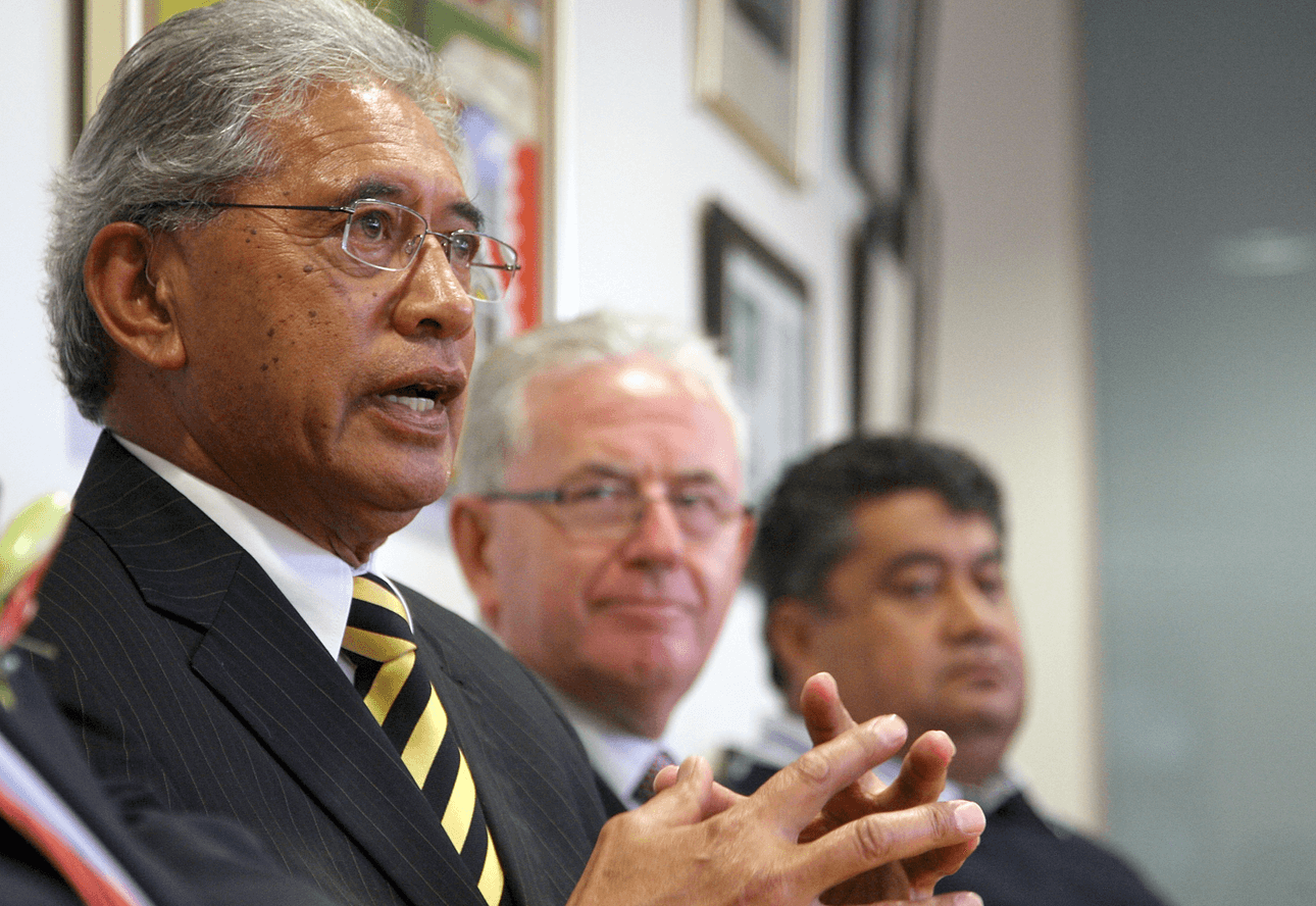 A man in a suit and striped tie speaks while gesturing with his hands. Two other men in suits sit beside him, listening attentively. Framed pictures are visible on the wall behind them.