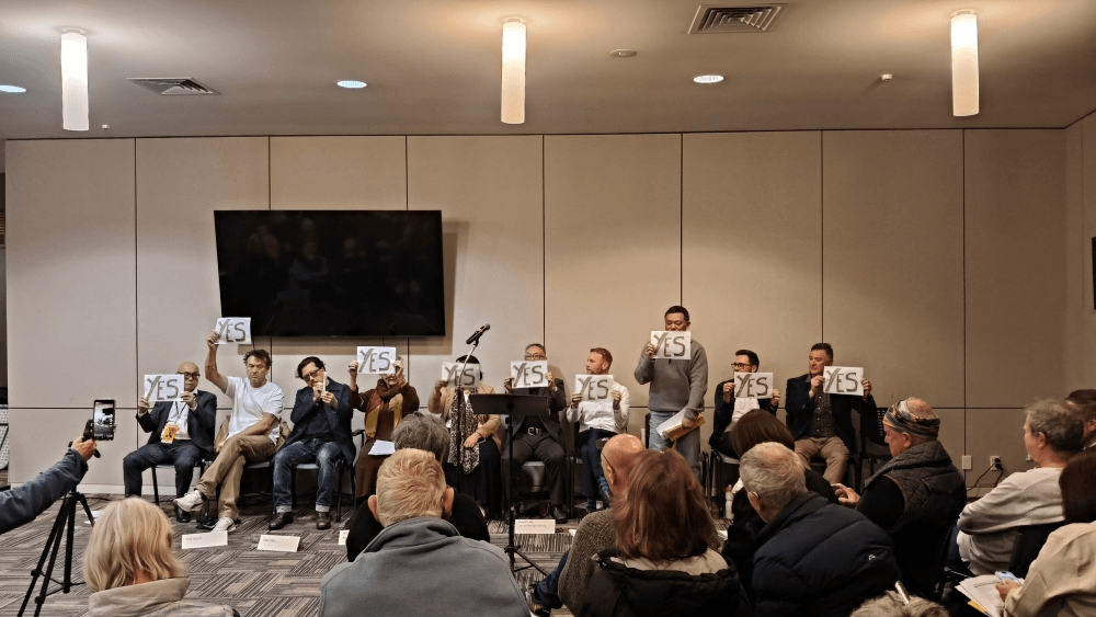 A panel of eight people sits at the front of a room holding signs that say "YES" while an audience watches. Microphones are on the table, and a large TV screen is mounted on the wall behind the panelists.