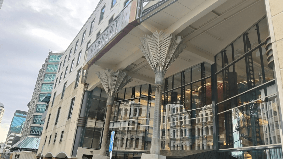 A modern library building with large glass windows reflecting historic architecture. Two metallic tree sculptures stand outside the entrance. Urban cityscape buildings are visible in the background under a cloudy sky.