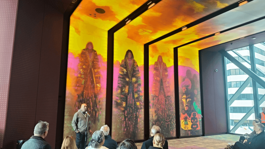 A man stands and speaks to a seated audience in front of a vibrant mural depicting four tall, cloaked figures and abstract, colorful designs on a large wall inside a modern building with floor-to-ceiling windows.