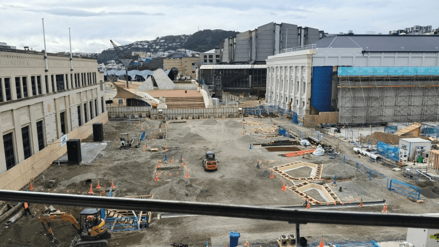 A large construction site with heavy machinery, scattered building materials, traffic cones, and fenced-off areas, surrounded by several large buildings under a cloudy sky.