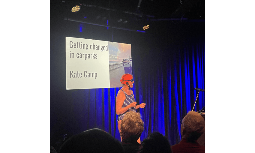 A stage with blue lighting. A screen reads "Getting changed in car parks". There is a woman, Kate Camp, standing in front of the screen wearing togs and a bathing cap.