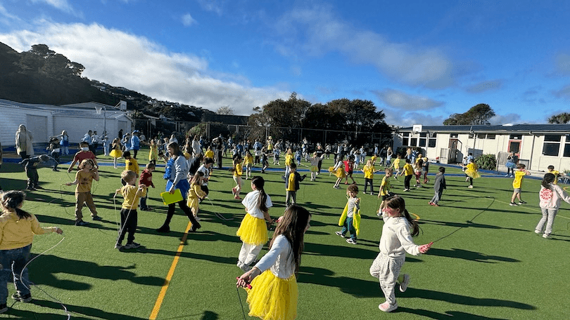 A large group of children, many wearing yellow, play and run around on a green outdoor sports field under a blue sky with scattered clouds. Adults and more children are visible in the background.