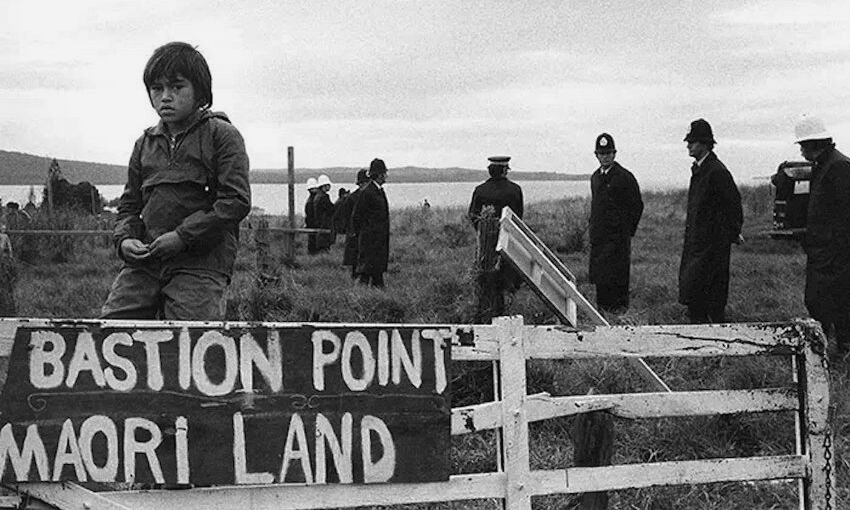 A young child sits on a broken fence with a sign reading "Bastion Point Maori Land." Police officers and other adults stand in the background on a grassy field near the water. The scene appears somber and tense.