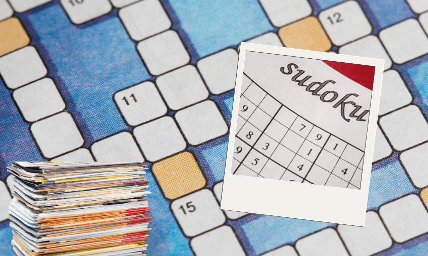 A close-up of a crossword puzzle grid with a stack of magazines in the bottom left corner and a polaroid photo of a completed sudoku puzzle placed on top of the grid.