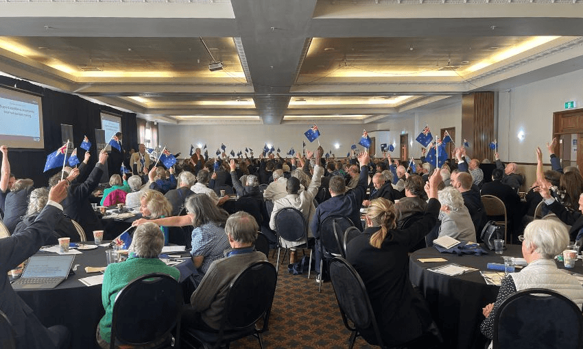A conference room filled with people sat at tables raise miniature New Zealand flags to raise a vote.