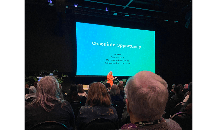 A photo of a large room full of people seated, with a stage and large screen. The screen is blue with white letters on it. There is a person dressed in pink in front of it.
