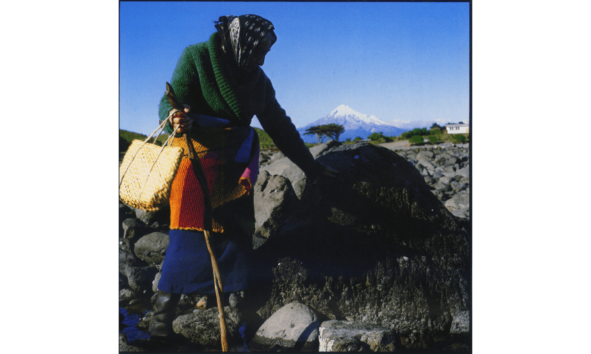 A colour photograph of a woman wearing woollen clothes and a headscarf, and holding a kete, and walking stick. She is outside and in a vast landscape with a mountain in the background. 