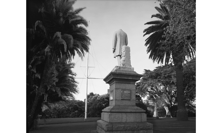 A black and white photo of an outdoor monument - the figure of a man - showing the figure's head has been chopped off. 