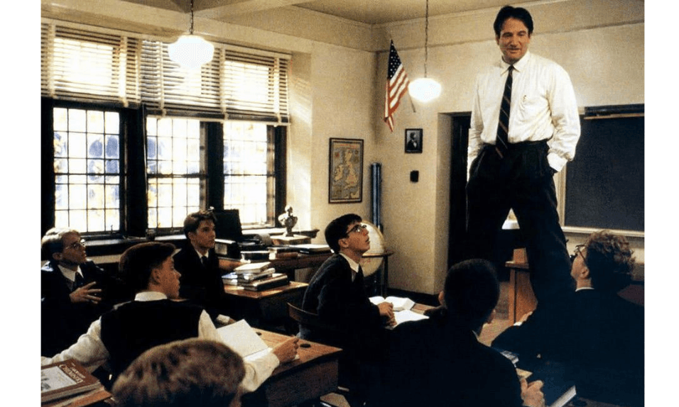 A film still from Dead Poets Society showing a classroom with boys seated at their desks while their teacher played by Robin Williams stands on top of a desk to address them. 