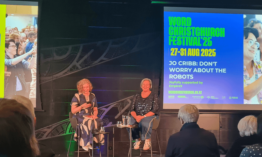 A photograph of an event at Word Christchurch with two women on a dark stage with a large screen behind them. The woman on the left is Susie Ferguson who is wearing a dress and has sort red hair. The woman on the right is Jo Cribb who is wearing pants and a top and has short brown hair.