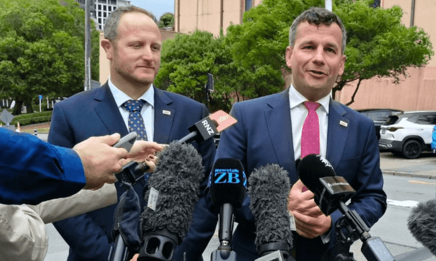 Two men in suits, the Act Party's Cameron Luxton and David Seymour, stand outdoors speaking at a press event. They are surrounded by microphones. Seymour is speaking, gesturing with his hand. Trees and a building are visible in the background.