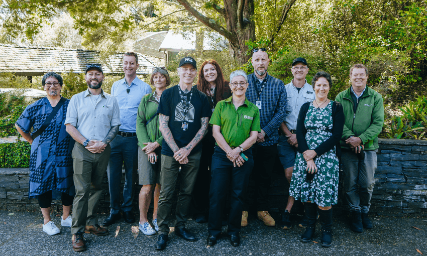 A group of twelve adults, some in uniforms and others in casual clothes, stand smiling outdoors in front of a stone wall and trees on a sunny day.