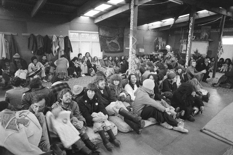A large group of people sit closely together on the floor of a dimly lit room, some wrapped in blankets and bundled in winter clothing, suggesting a communal gathering or meeting.