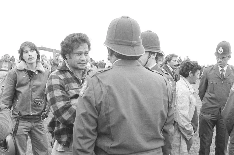 A group of people, including protesters and uniformed police officers, stand facing each other outdoors. The scene appears tense, with onlookers in the background. The photo is in black and white.