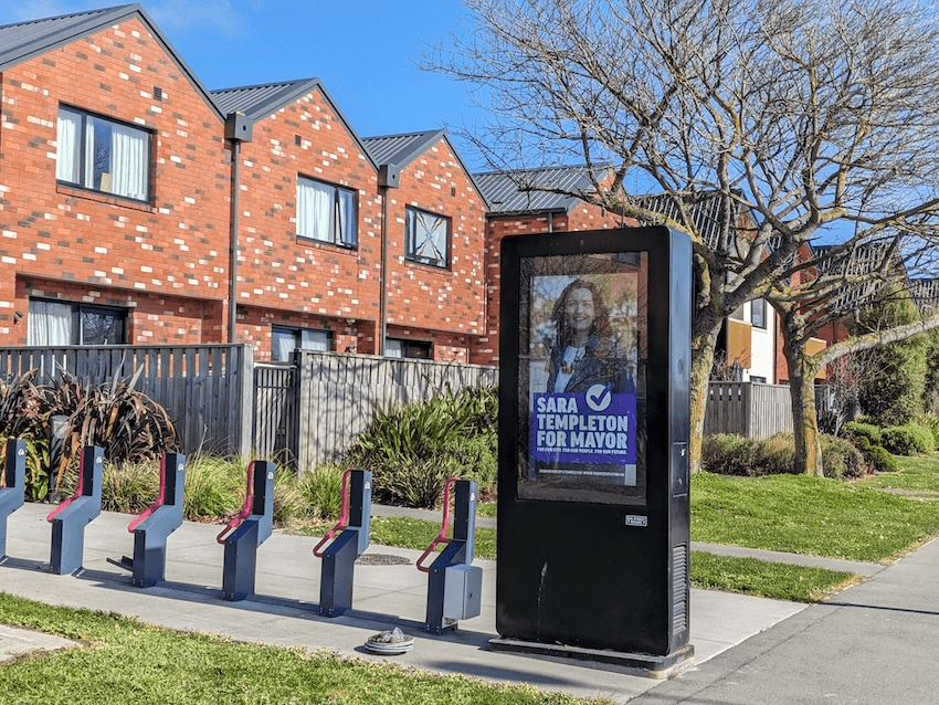 a sunny street where there are some brick housing in the background and a digital billboard of a pakeha woman with a purple jacket reading 'sara templeton for mayor'
