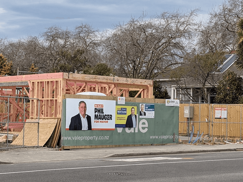a timber frame of a house being built and a 're-elect Phil Mauger sign in the foreground with shades of orange, and a hanful of other centre right candidate posters at a smaller size