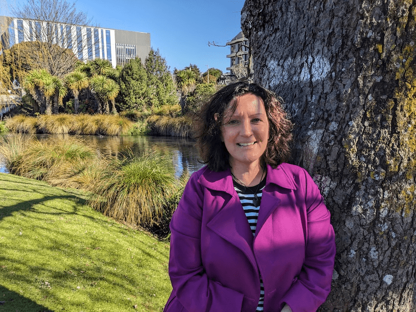 Sara, a woman in a purple coat, smiles next to a tree in the sunshine with a river in the background
