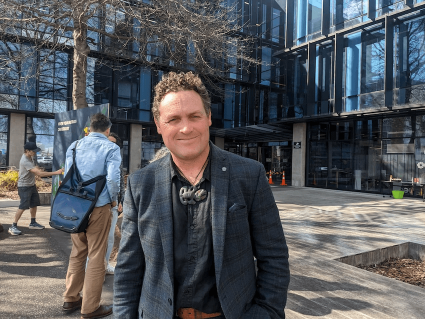 Craig pauling, a middleaged man wearing a large pounamu pendant, smiles in the sunshine with the ECan building behind him