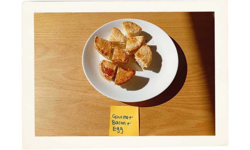 A white plate with six pieces of savory pastry sits on a wooden surface. Below the plate is a yellow sticky note labeled "Gourmet Bacon + Egg" handwritten in blue ink.
