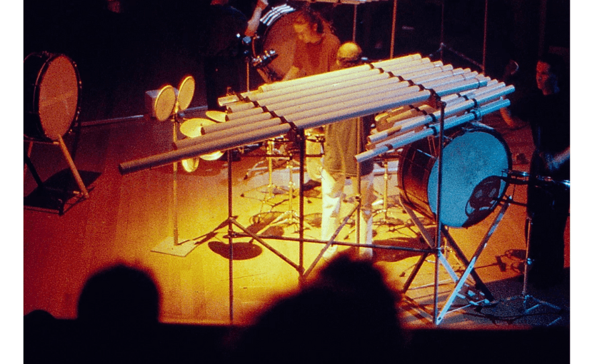 A blurry stage with large instruments set up. There are three musicians on stage and several percussion instruments.