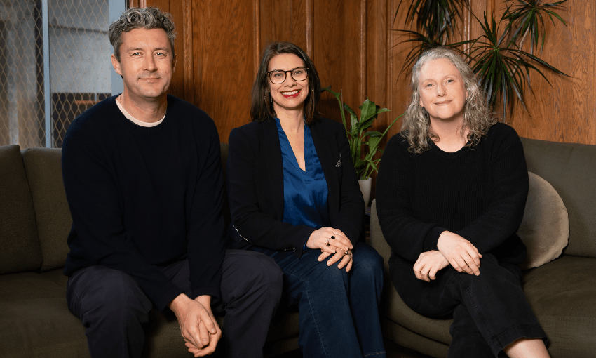 three people smiling while sitting on a couch and wearing dark clothing. one is a man with salt and pepper hair, one is a woman with dark glasses and dark hair and the other is a woman with curly grey hair