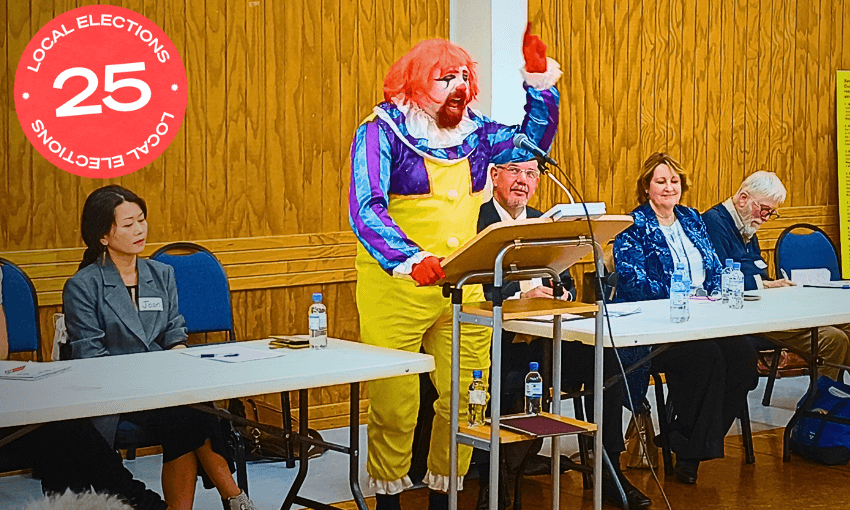 A person dressed as a clown with bright red hair, yellow overalls, and colorful makeup speaks energetically at a podium, while four seated adults in business attire look on in a wood-paneled room.
