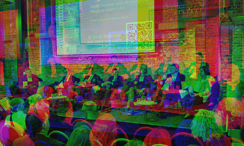 A panel of speakers sits on stage in front of an audience, with a large screen displaying event information and a QR code on a brick wall behind them. The event appears to be a discussion or forum.