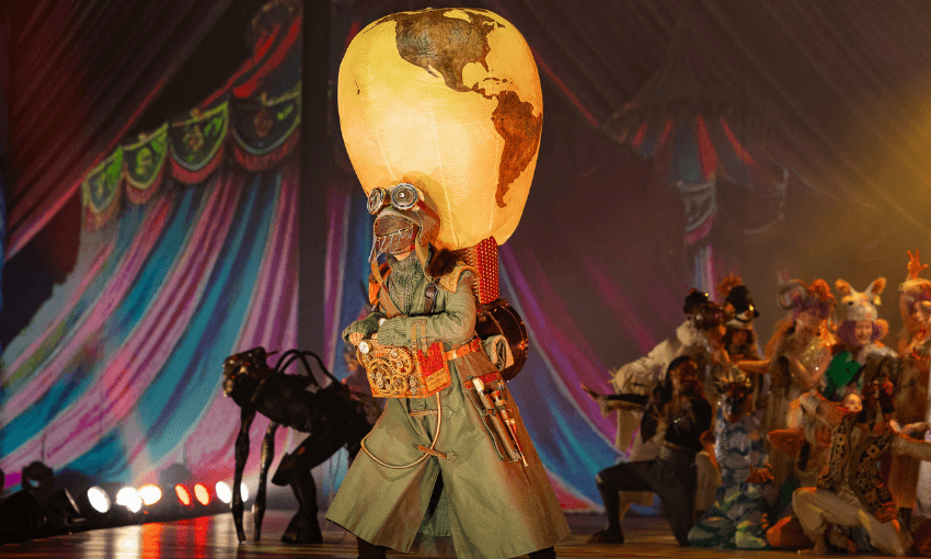 A performer in a long coat and goggles wears a large globe-shaped balloon helmet on stage, surrounded by other colorful, elaborately costumed actors under circus-like lighting and draped decorations.