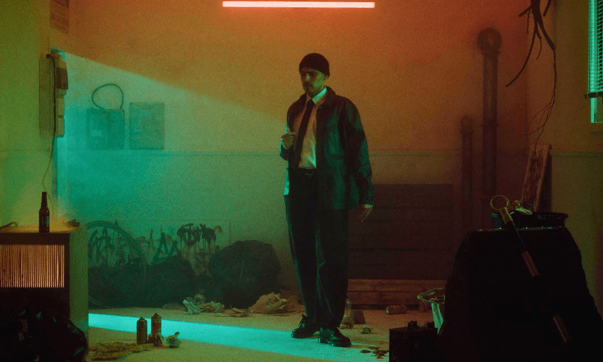A man in a black jacket and beanie stands in a dimly lit, cluttered room with neon and green lighting. The floor is scattered with trash, and the atmosphere is moody and mysterious.