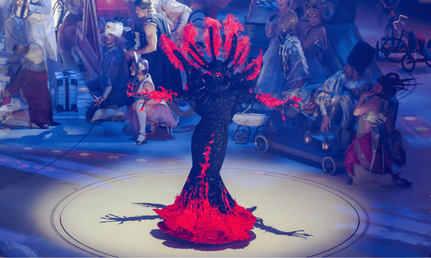 A performer in a black costume with dramatic red feathers extending from their arms and head stands center stage, surrounded by other costumed performers in an elaborate theatrical or circus setting.