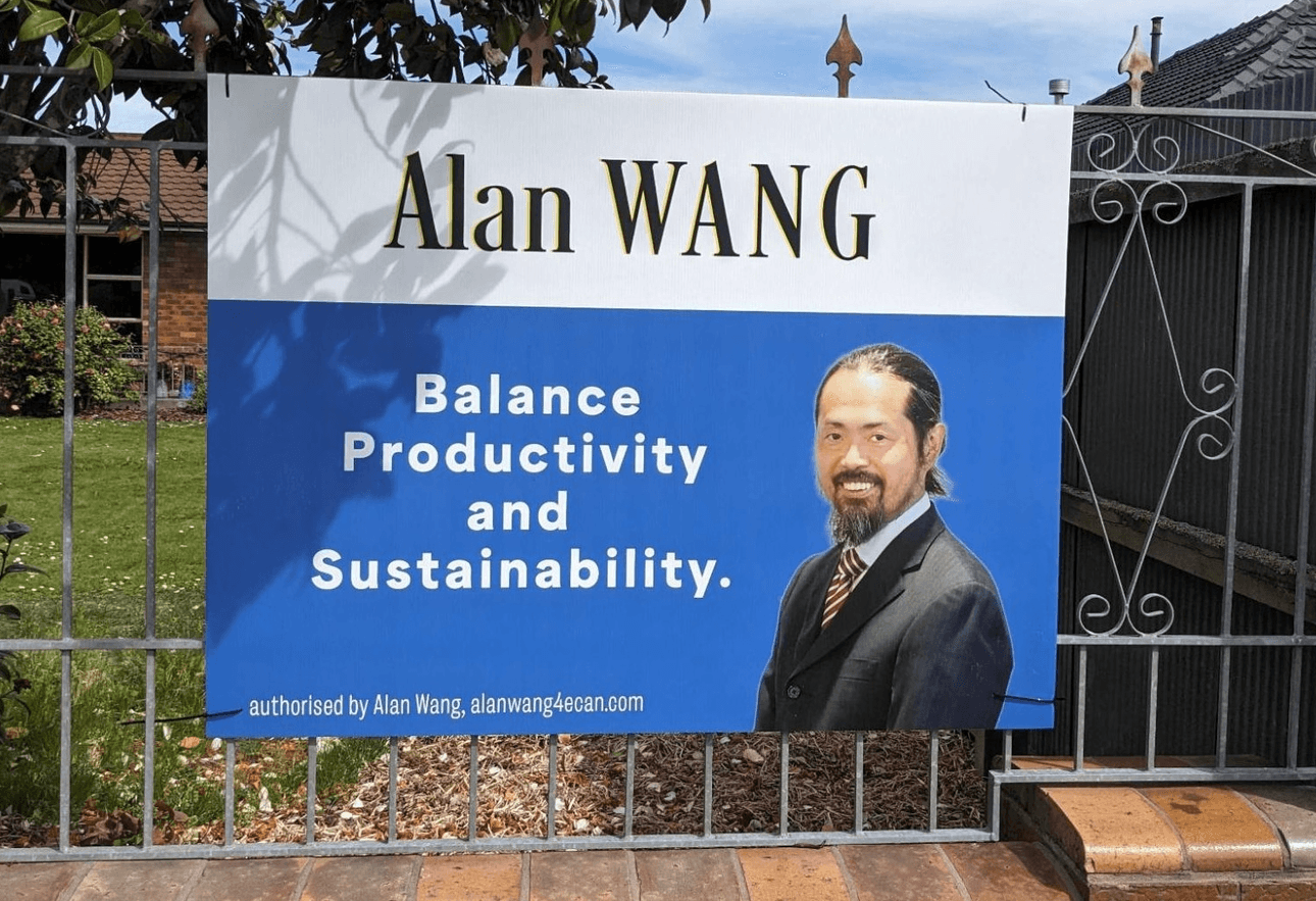 A campaign sign on a metal fence reads “Alan WANG. Balance Productivity and Sustainability,” with a photo of a smiling man in a suit and tie. Lush greenery and a house are visible in the background.