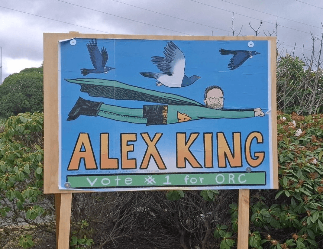 A campaign sign shows a cartoon of a caped person flying among birds, with the name "ALEX KING" in large letters and the text "Vote #1 for ORC" below. The sign is outdoors, surrounded by bushes.