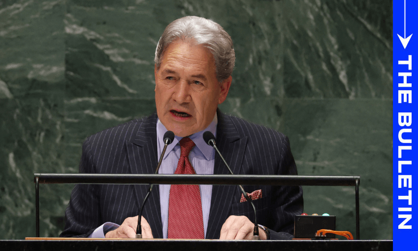 Winston Peters wears a pinstripe suoit and red tie while speaking at a lectern. A green marble wall is behind him