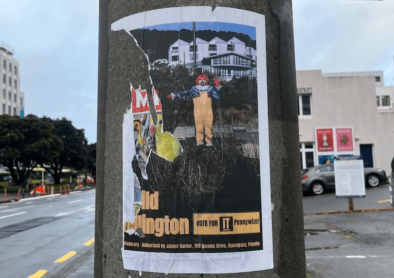 A torn poster on a city lamppost shows a clown in front of houses, with partially visible text: "Vote for IT Pennywise" and some address details. Buildings, trees, and a wet street are in the background.